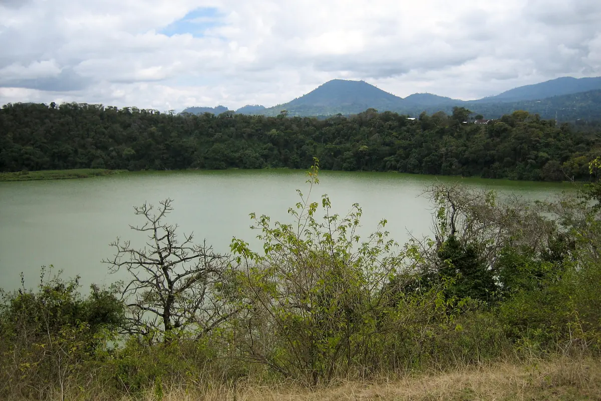 Ruhiger grüner Kratersee mit bewaldetem Ufer und Hügeln dahinter